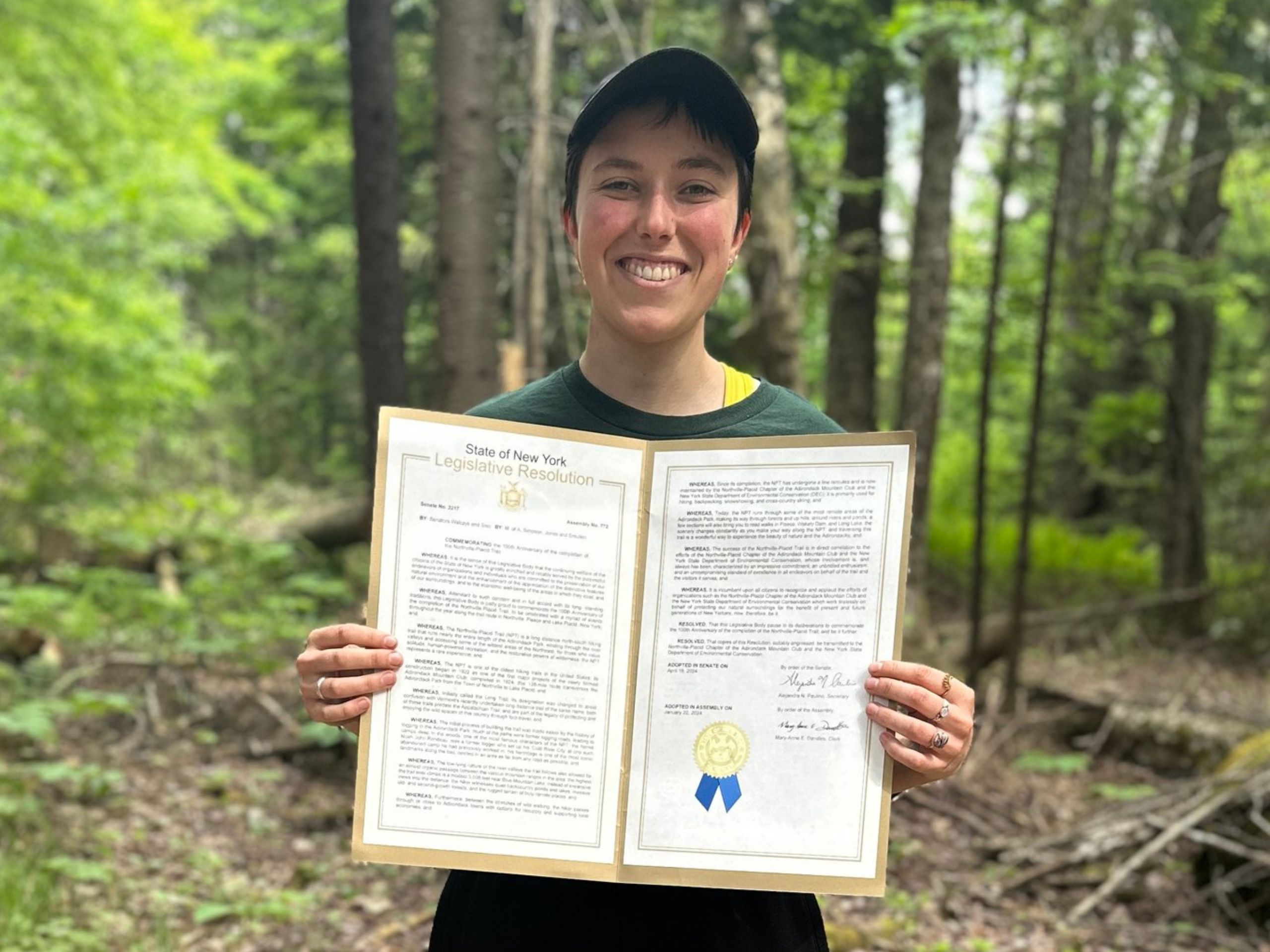 a woman wearing a hat while standing in the woods and holding a proclamation