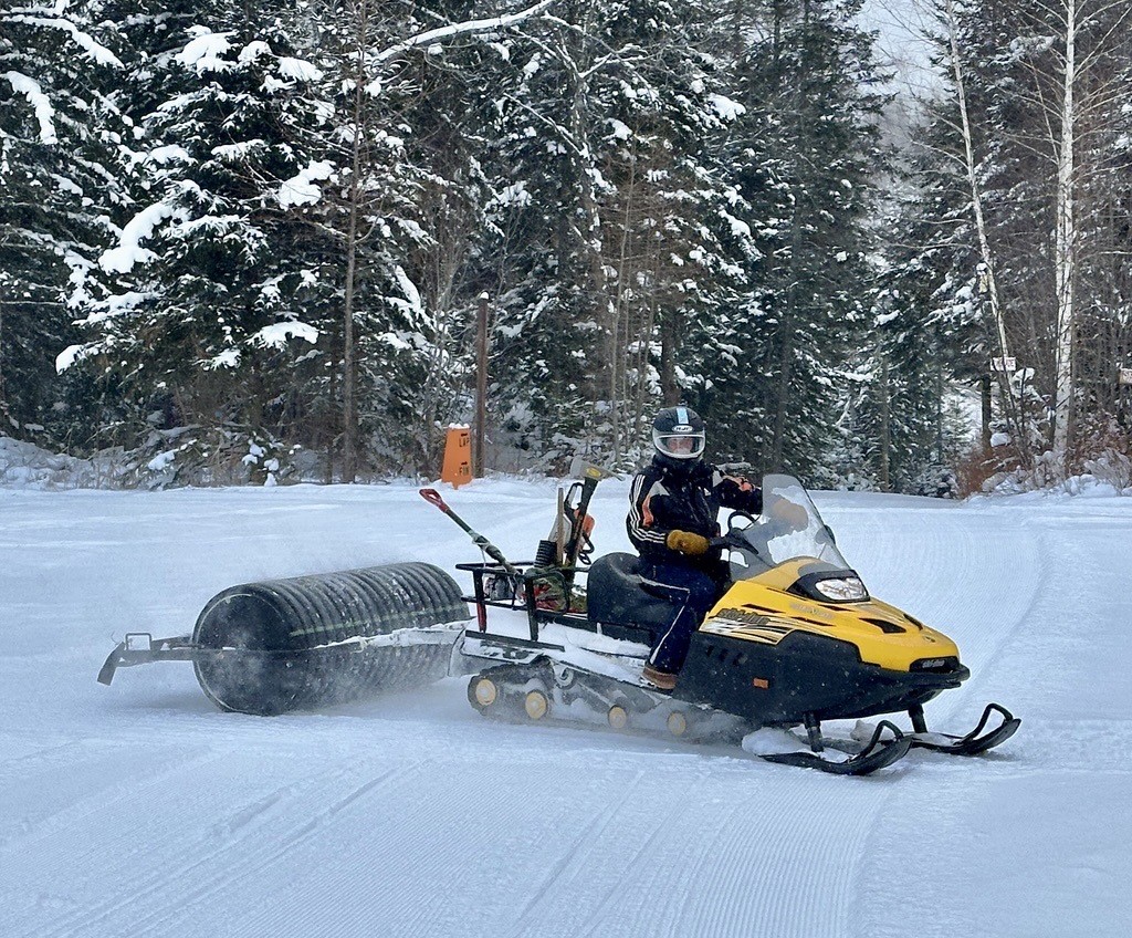 man riding a yellow snowmobile with trail grooming supplies in the back