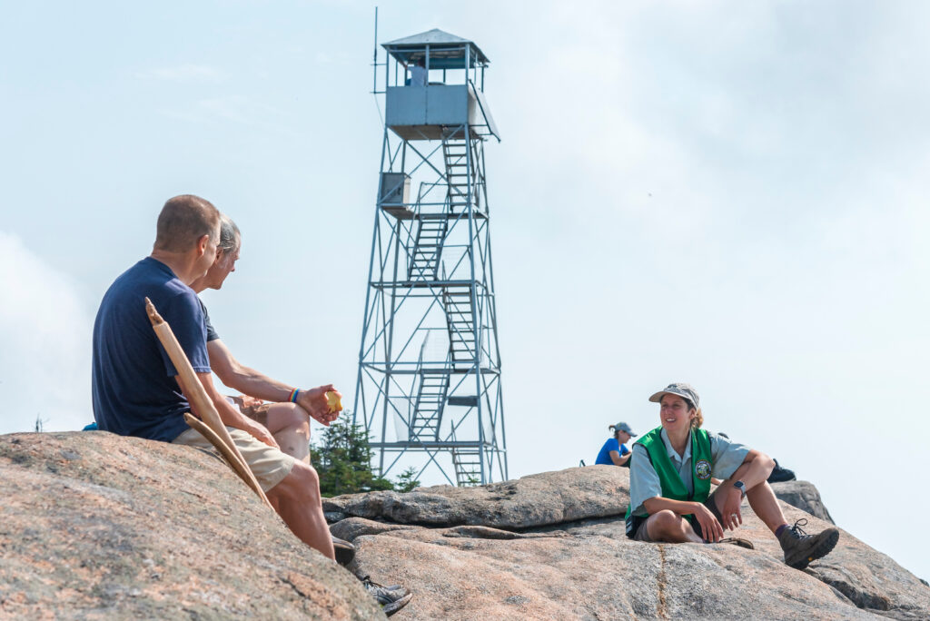a summit steward speaks to hikers on a mountain summit with a fire tower in the background