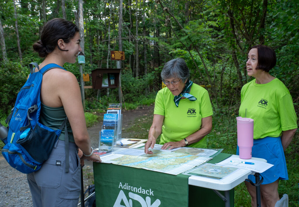 two trailhead stewards in bright green shirts speak to a hiker at the Buck Mountain-Pilot Knob trailhead