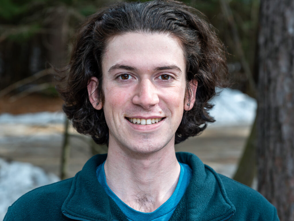 person with long dark hair stands in front of wooded background