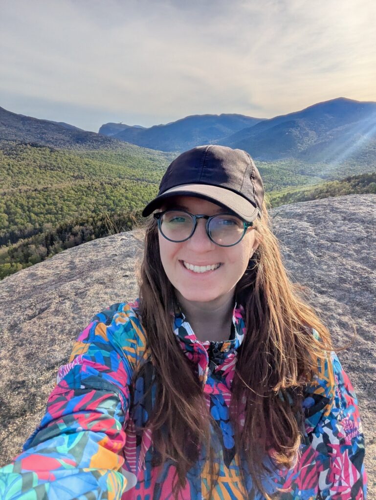selfie of a woman wearing a hat and glasses at the summit of a mountain