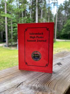 Red Adirondack High Peaks Summit Journal resting on a weathered wooden railing with a green forest backdrop.