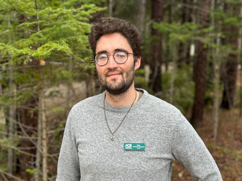 man wearing glasses, a necklace, and gray sweater posing for a picture with a wooded background