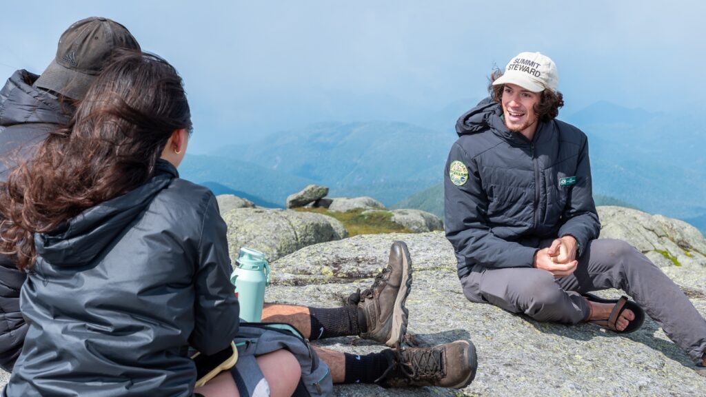 summit steward wearing a hat and jacket speaks to two hikers