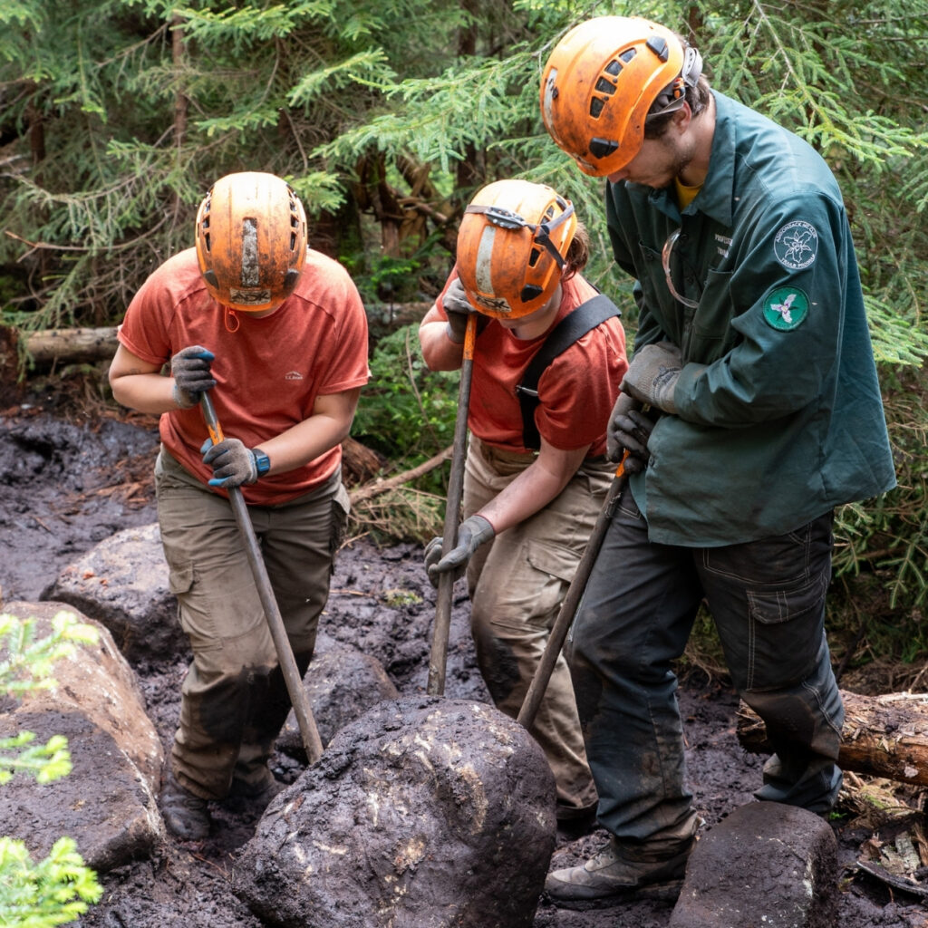 three trail crew workers using rock bars to move a rock