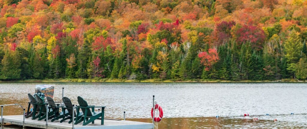 Adirondack chairs on a dock on a lake in autumn with foliage in the background