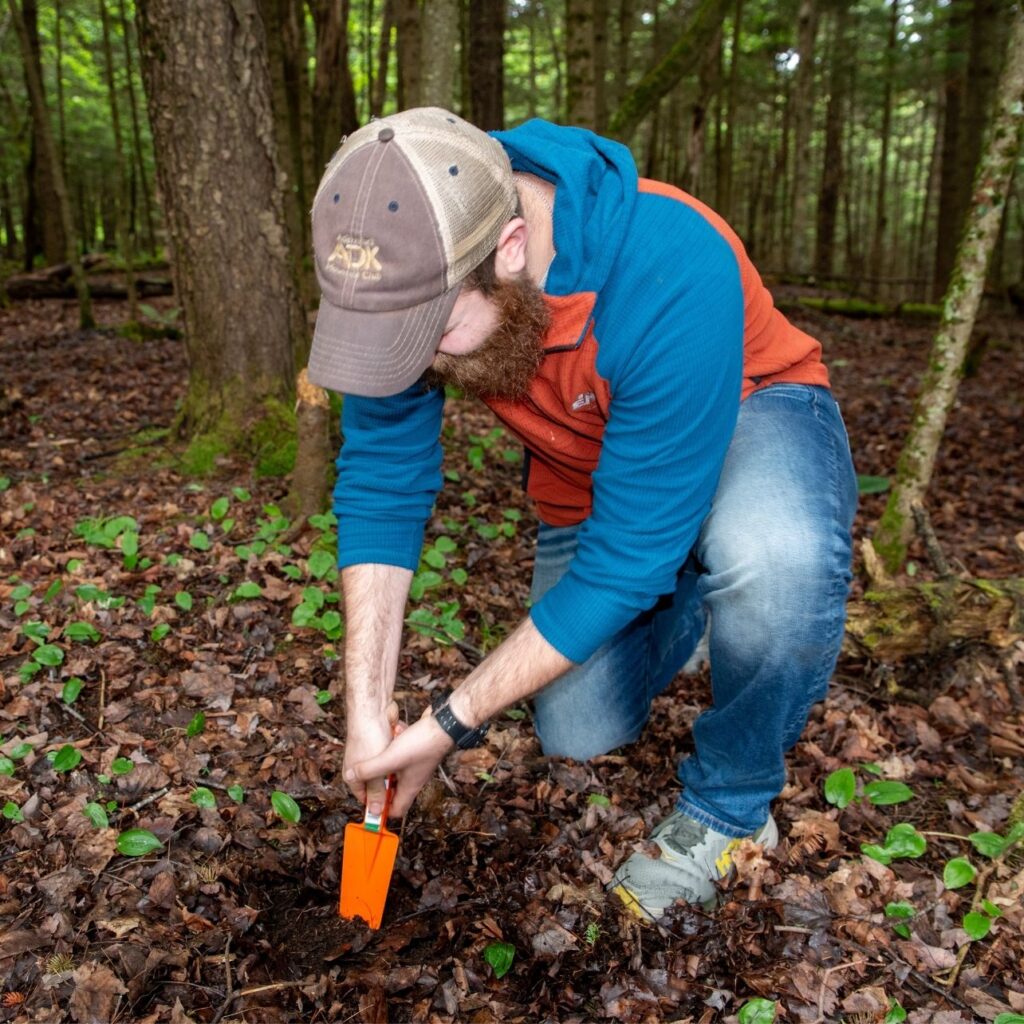 man wearing a hat digs cathole in woods
